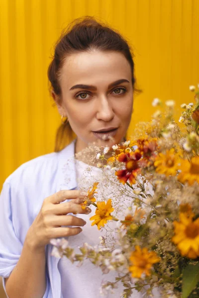 Güzel esmer kadınla sarı arka plan üzerinde topuz kafasındaki alanları çiçekler elinde tutarken mavi gömlek ve beyaz tişört giyiyor. Erkek çiçek getirmek — Stok fotoğraf