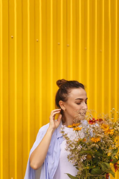 Mulher morena bonita no fundo amarelo com coque na cabeça vestindo camisa azul e camiseta branca enquanto segurando campos flores na mão. Feminino trazendo a flor — Fotografia de Stock