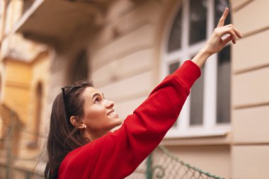 güzel mimari ile sokakta yürüyen mutlu turist kadın. esmer kız bir şey arıyor. Bazı binasında parmakla işaret eden bir kadın. o kırmızı kapüşonlu giyiyor.