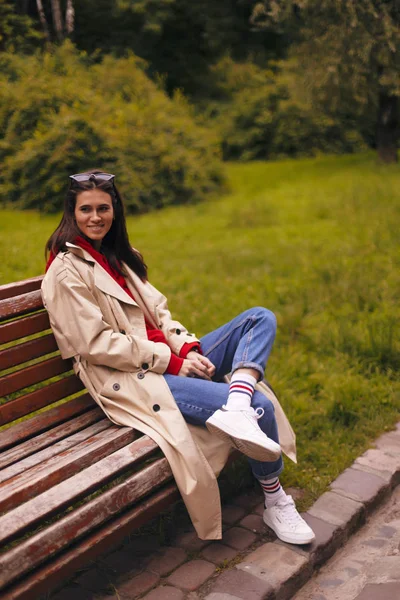 Porträt einer Frau in voller Länge mit beigem Trenchcoat, rotem Kapuzenpulli, blauer Jeans und weißen Turnschuhen beim Spazierengehen im Park. — Stockfoto