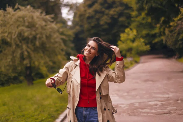 Porträt einer Frau in beigem Trenchcoat und rotem Kapuzenpulli beim Spazierengehen im Park. sie sieht glücklich aus. — Stockfoto