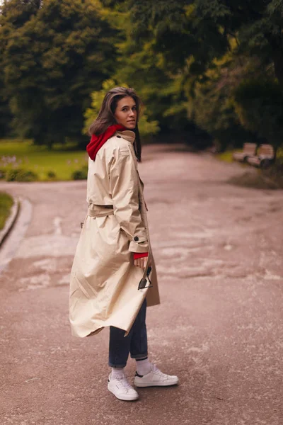 Retrato de mujer de cuerpo entero con gabardina beige, sudadera con capucha roja, vaqueros azules y zapatillas blancas mientras camina por el parque . —  Fotos de Stock