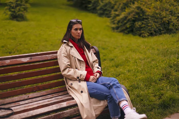 Porträt einer Frau in voller Länge mit beigem Trenchcoat, rotem Kapuzenpulli, blauer Jeans beim Spazierengehen im Park und auf einer Bank sitzend. — Stockfoto