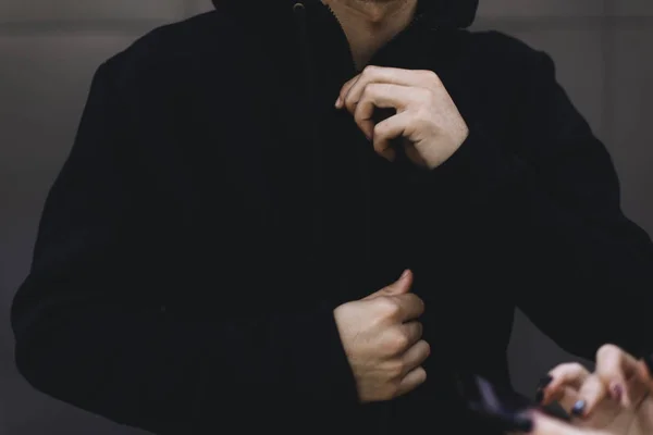 Young man zipping up his hoodie. Man dressing black sport clothes. Grey urban background. In the foreground are women\'s hands with a telephone.