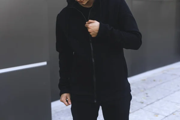 Tânărul şi-a zipat hanoracul. Bărbat îmbrăcat haine sport negru. Fundal urban gri . — Fotografie, imagine de stoc