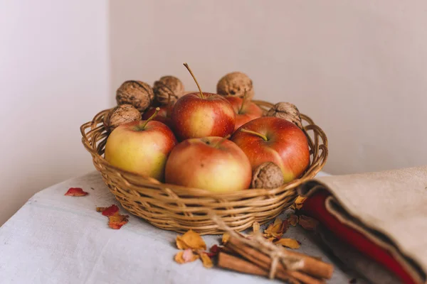 Still life with apples in a wicker basket, cloth, walnuts, flower petals and cinnamon sticks wrapped in twine. Concept of fall or winter home comfort. — Stock Photo, Image