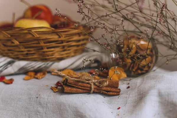 Still life with cinnamon sticks wrapped in twine, apples in a wicker basket stand on a striped linen cloth, walnuts and flower petals. Concept of home comfort in autumn or winter. — Stock Photo, Image