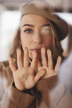 Cam pencere, kapak ağız altına hayran güzel kadın bakın. Kadın toplam göz giyer. Bej renkli bere. Cama el.