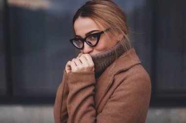 İş Merkezi windows yürüme çekici kadın. Sarışın kadın model fotoğrafı kapak ağzı ile kazak boyun tarafından görünüyor. Sıcak ve konfor kavramını. Kız giyim gözlük, ceket, boyunlu kazak. 