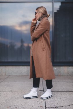 Çekici Kıvırcık sarışın kadın ceket, beyaz spor ayakkabı, siyah kot pantolon ve kazak giymiş. İş merkezinin arka planda pencereler. Sokak moda bahar, kış ve sonbahar sezonu için. Minimalist bak. 