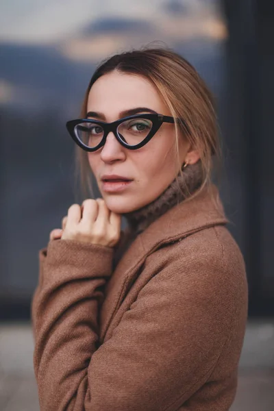 Attractive beauty woman walking near business center while wearing beige coat and glasses. Photo blonde girl which turn around. The window is a reflection of the sky