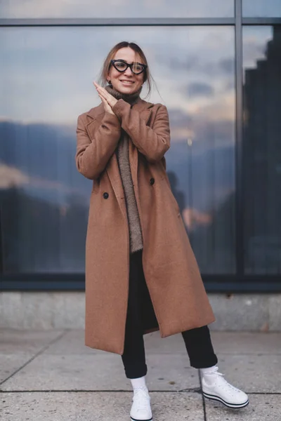 Attraktive blondgelockte Frau mit Mantel, weißen Turnschuhen, schwarzen Jeans und Sweatshirt. Hintergrund Fenster des Business Centers. Straßenmode für Frühling, Winter oder Herbst. minimalistischer Look. — Stockfoto