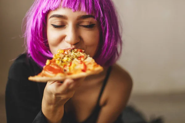 Grappige roze haar pruik meisje in zwarte lingerie en shirt eten pizza thuis. Meisje genieten van een heerlijke pizza. — Stockfoto