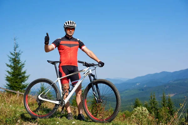 Happy professional sportsman cyclist standing with cross country bicycle on hill and raising hand up against blue sky with clouds at summer sunny day, Outdoor sport, success and life goals concept