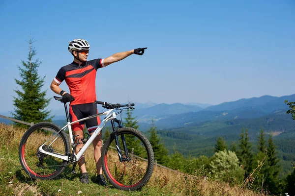 Профессиональный Велосипедист Спортивной Одежде Шлеме Стоящий Вершине Холма Указывающий Голубое — стоковое фото
