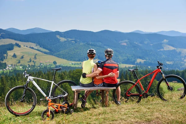 Young family tourists cyclists resting on wooden bench with bikes on grassy hill with distant mountains view background, Active lifestyle and happy relations concept