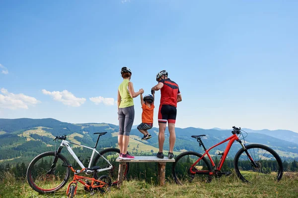 Young family tourists cyclists resting on wooden bench with bikes on grassy hill with distant mountains view background, Active lifestyle and happy relations concept
