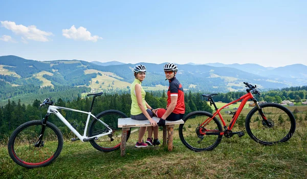 Happy couple sportsman cyclists sitting on wooden bench near bicycles and looking at camera while resting after riding bikes in mountains, Active lifestyle, traveling and outdoor sport concept