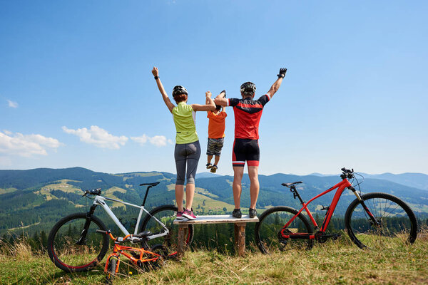 Young family tourists cyclists resting on wooden bench with bikes on grassy hill with distant mountains view background, Active lifestyle and happy relations concept 