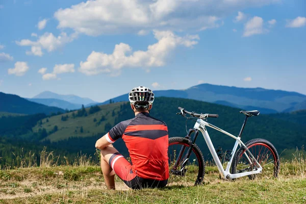年轻的运动员骑自行车在专业运动服和头盔坐在单车附近的草路边在阳光明媚的一天 积极的生活方式和户外运动的概念 — 图库照片