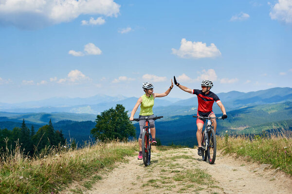 active couple cyclists in professional sportswear and helmets giving each other high five while  cycling down cross country bikes on  mountain road at summer day, Active lifestyle and outdoor sport concept