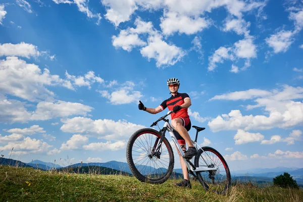 Happy professional sportsman cyclist standing with cross country bicycle on hill and showing thumb up against blue sky with clouds at summer sunny day, Outdoor sport, success and life goals concept