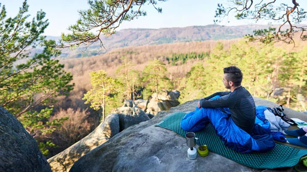 Young bearded handsome man sitting alone in sleeping bag on big rock and having coffee while enjoying beautiful mountain view on clear sunny summer morning, Tourism and active lifestyle concept