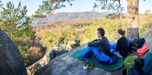 Young tourist couple sitting in sleeping bags on big rock and enjoying magnificent peaceful view of mountains covered with forest on early morning, Active lifestyle concept