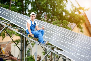 Neredeyse oturan genç gülümseyen elektrikçi tek başına güneş Fotoğraf volta panel sistemi tornavida ile jest yukarı parlak güneşli yeşil ağaç arka plan üzerinde gösterilen bitmiş