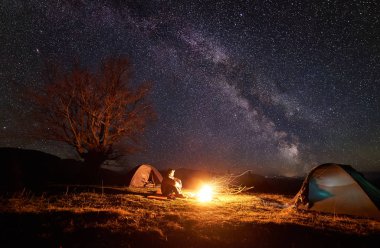 Dağda kamp. İki çadır kamp ateşi güzel koyu mavi yıldızlı gökyüzü arka plan üzerinde parlakça yanıyor çimenli Vadisi izlerken arasında genç turist adam oturur. Turizm ve doğa kavramının güzellik.