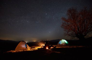 Kamp gece dağlarda. İki genç turistler, erkek ve kız arasında iki çadır kamp ateşi arka plan koyu yıldızlı gökyüzü altında yanan üzerinde arka arkaya oturan siluetleri. Turizm ve seyahat kavramı.