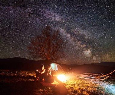 Kamp gece dağlarda. Karanlık siluetleri iki genç turistler, oturma ve kız çadır ve uzak tepelerin arka plan üzerinde koyu yıldızlı gökyüzü altında kamp ateşi yakarak yaktı. Turizm ve seyahat kavramı.