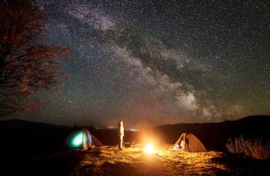 Dağlarda huzurlu kamp gece. Genç turizm uzun saçlı kız çok parlak parlak yıldız ile derin karanlık gökyüzü izlerken kamp ateşi yakma adlı iki çadır arasında duruyor. Turizm ve seyahat kavramı.