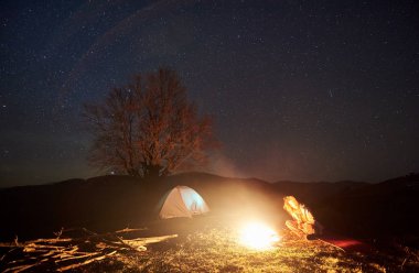 Kamp gece dağlarda. Genç kadın backpacker derin karanlık yıldızlı gökyüzü altında kamp ateşi yakma yakınındaki bir dinlenme. Parlayan çadır ve büyük ağaç arka planda silüeti. Turizm ve seyahat kavramı.