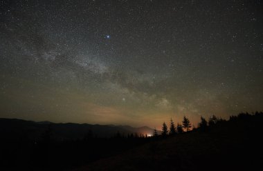 Fantastik dağlar geceleri görüntüleyin. Sarp tepeler köknar ağaçlarının silhouettes parlak parlayan yıldızlar ve ufuk uzak ışık ile karanlık mavi gökyüzü karşı orman ile kaplı. Doğa kavramının güzellik.