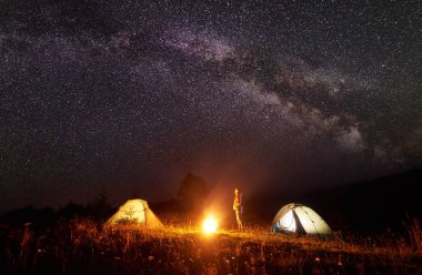 Dağda kamp. Genç turist kız iki çadır ile parlak parlayan yıldızlar arka plan çok derin, karanlık gökyüzü parlak yanan ateşin ortasına izlerken arasında ayakta ince. Turizm ve seyahat kavramı.