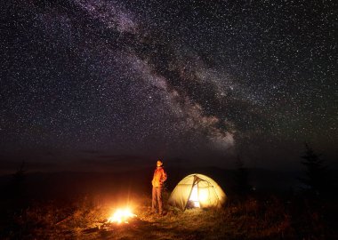 Gece dağda kamp. Işıklı çadır dinlenme ve parlak parlak parlayan yıldız ve Samanyolu çok derin, karanlık gökyüzü ateşin ortasına yanan kadın. Turizm ve açık hava etkinliği konsepti.