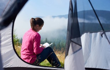 Genç uzun saçlı turist kız pembe kazak ve küçük çadır girişte oturma ve parlak yaz sabahı güzel sisli dağlar arka plan üzerine bir kitap okuma kot yakın çekim portre.