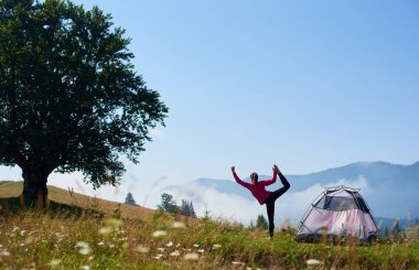 Yoga poz turist çadır ve büyük ağaç sisli dağlar arka plan üzerinde güzel mavi gökyüzünün altında yeşil çimenli tepe üzerinde tek ayak üzerinde duran genç ince kız arkadan görünüm. Turizm ve kamp kavramı.
