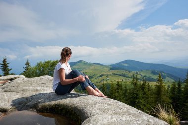 Profil görüntüleme genç uzun saçlı yalınayak ayakkabın çam ormanı ile kaplı Dağları'nın muhteşem manzarayı güzel mavi gökyüzü altında orta ile büyük kayanın üzerine oturan turist kız.