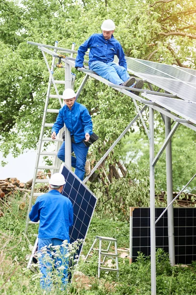 Installing of stand-alone solar photo voltaic panel system. Workers in hard-hats and blue overall lifting the solar module on metal platform. Alternative energy and professional construction concept.