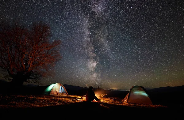 Gece dağda kamp. İki ışıklı çadır, kamp ateşi, güzel yıldızlı gökyüzü ve Samanyolu arka plan üzerinde zevk arasında oturan erkek uzun yürüyüşe çıkan kimse silüeti. Turizm, açık hava etkinliği konsepti