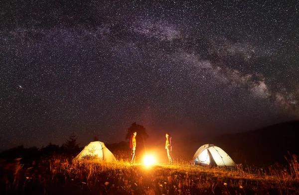 Night camping. Bright campfire burning between two hikers, man and woman standing opposite each other in front of shining tents under amazing dark starry sky with Milky way on distant hills background