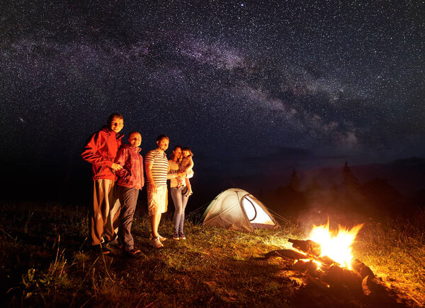Night camping in mountains. People standing in front of burning campfire. Illuminated tent and starry sky and Milky way on background. Mother holding in arms daughter. Tourism and recreation concept.