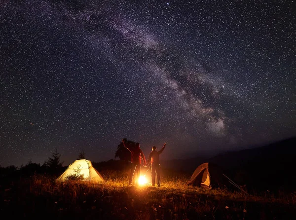 Night camping in the mountains. Bright campfire burning between two silhouettes of tourists man and woman standing with arms raised up in front of tents under beautiful dark starry sky and Milky way