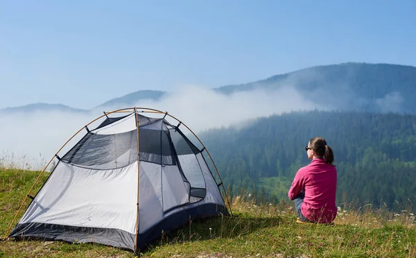 Arkadan görünüşü genç uzun yürüyüşe çıkan kimse kadın turist çadır, çiçek açan Vadisi'nin yeşil çimenlerin üzerinde oturan güneş gözlüğü, güzel mavi gökyüzü zevk, sisli dağlar parlak yaz sabahı görüntüleyin.
