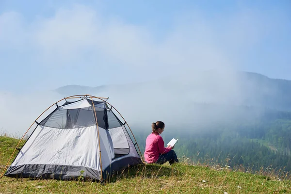 Arkadan görünüşü turist çadır sisli dağlar arka plan üzerinde parlak yaz sabahı bir kitap okuma güzel mavi gökyüzünün altında çiçek açan Vadisi'nin yeşil çimenlerin üzerinde oturan genç uzun saçlı turist kız.