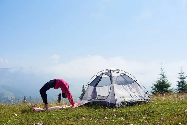 Genç kız üzerinde parlak güneşli sabah küçük turistik çadır altında güzel mavi gökyüzü, yeşil çimenlerin üzerinde jimnastik egzersizleri yapıyor yalınayak ince. Spor, turizm ve kavram kamp dağ.