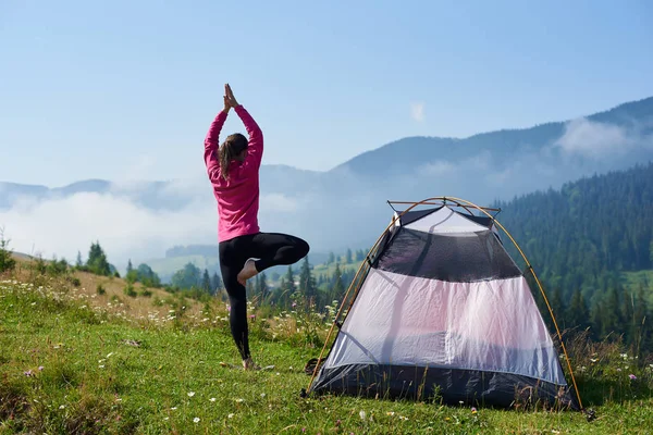Back view of young long-haired barefooted tourist girl standing on one leg in yoga pose with raised arms on green grassy valley at tourist tent under beautiful blue sky on foggy mountains background.