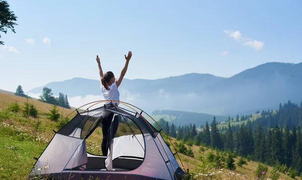 Genç mutlu kadın ile ayakta arkadan görünüm parlak yaz sabahı çiçek açan hill adlı küçük turistik çadır güzel berrak mavi gökyüzünün altında silah kaldırdı. Turizm ve kamp kavramı.